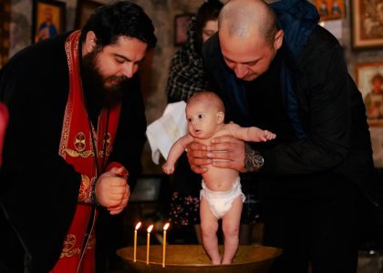 A priest and father performing an Orthodox baptism ceremony with a baby.
